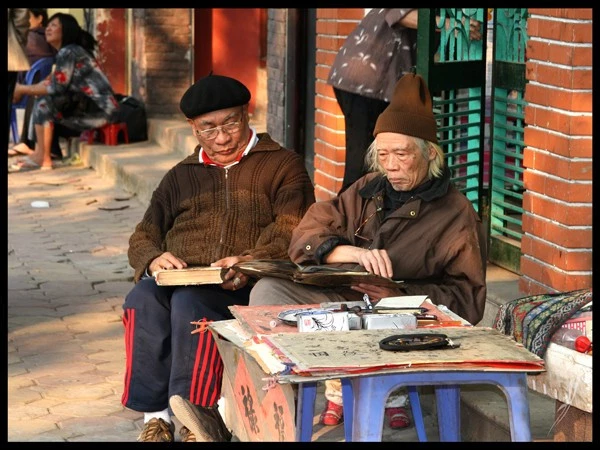 Cuối năm 2010, Hà Nội vẫn còn thở theo nhịp chậm, nơi vỉa hè không chỉ để đi mà còn là nơi hành nghề, trao chuyện, và lắng ký ức. Hai người đàn ông ngồi ngồi đó, không cần lời, như một phần của phố, của thời gian, của một lớp văn hóa đang lặng lẽ bền bỉ tồn tại giữa đổi thay.