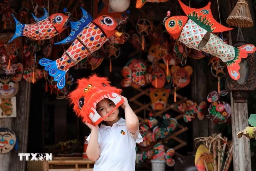 Em nhỏ thích thú đội đầu lân giữa không gian rực rỡ của đèn cá chép, mặt nạ và đồ chơi Trung thu truyền thống. (Ảnh: Phạm Tuấn Anh/TTXVN)